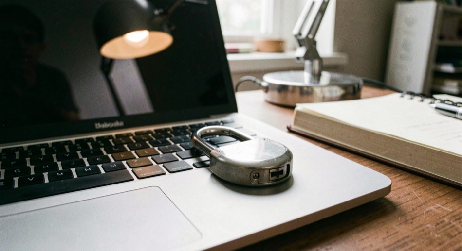 A closed padlock resting on a laptop keyboard next to a notebook, the screen dark and a desk lamp reflected across its surface